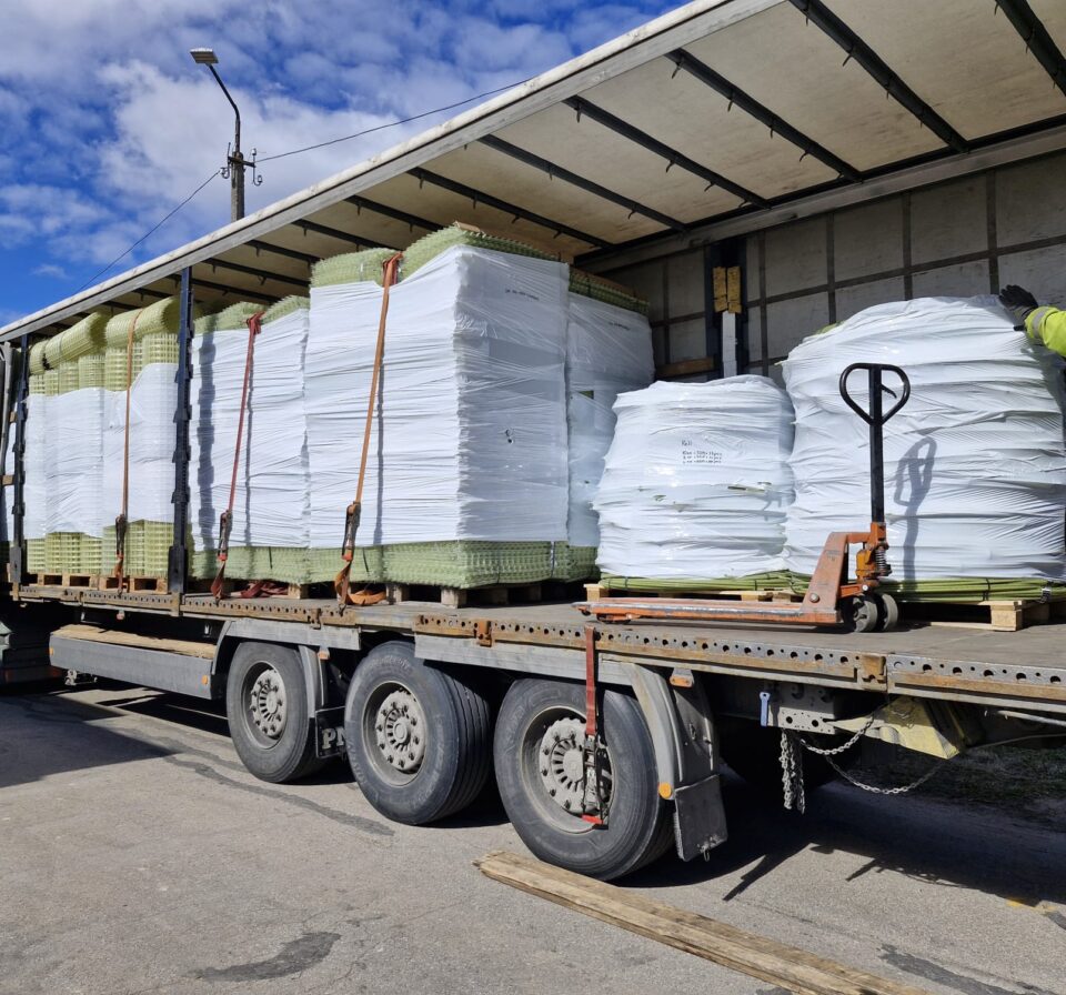 Truck loaded with palletized GFRP mesh and composite rebars secured with straps, ready for transport. Fiberglass reinforcement materials wrapped in protective film demonstrate organized logistics and efficient construction material delivery.